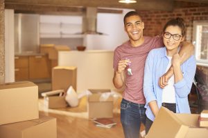 a young couple unpack their belongings as they settle into their new loft apartment . They are hugging and looking to camera smiling holding their keys aloft .
